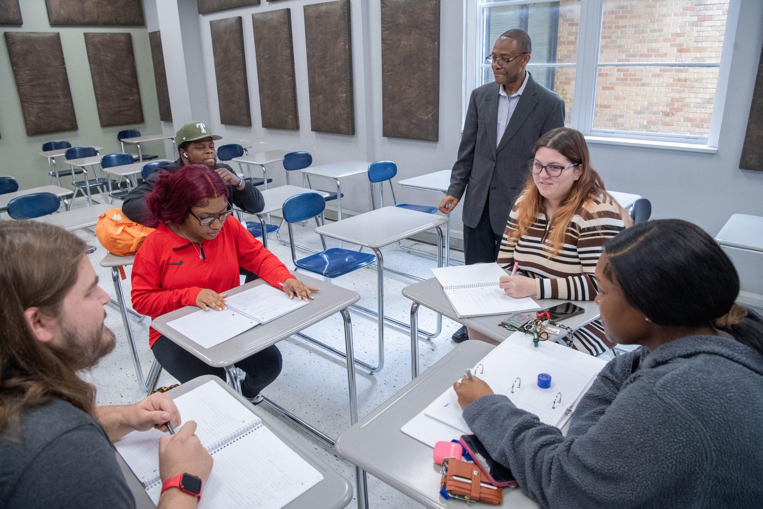 五个学生拿着打开的笔记本，一组坐在教室里，旁边站着一位教授.